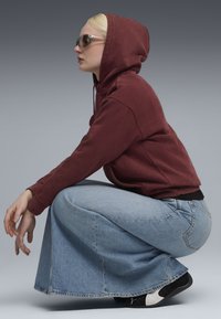 Person iført en bordeaux hættetrøje, lysblå denimskørt, sort og hvidt sneakers og stjernesolbriller, sammenkrøbet mod en grå baggrund.