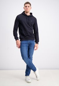 Marineblauer Hoodie mit einer vorderen Tasche, Kapuze mit Kordelzug und gerippten Bündchen, kombiniert mit blauen Jeans und weißen Sneakern vor einem schlichten Hintergrund.