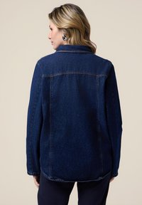 Jachetă din denim de culoare albastru închis, cu guler clasic, mâneci lungi și tiv curbat. Cosea vizibile și croială lejeră.