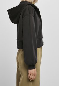 Sudadera negra recortada con cierre de cremallera, capucha extragrande y puños elásticos. Tejido suave con textura lisa, combinada con pantalones beige.