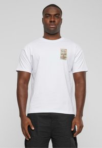 Hvid bomulds T-shirt med korte ærmer, der har et lille multifarvet grafisk mærke på venstre bryst, som viser tekst og designelementer.