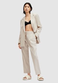 Completo in lino beige con giacca su misura e pantaloni a vita alta, abbinato a un reggiseno nero e sandali bianchi. Tessuto morbido, vestibilità comoda.