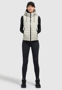 Hellgraue gepolsterte Weste mit Kapuze, Front-Reißverschluss und Seitentaschen. Trägt man über einem schwarzen Langarmoberteil und kombiniert mit schwarzen Leggings und Schuhen.