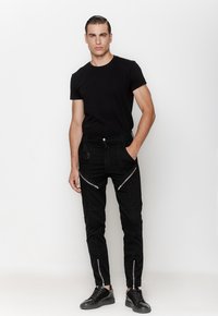 Czarny dopasowany t-shirt w połączeniu z czarnymi dżinsami z wieloma zasuwanymi kieszeniami i teksturowanymi panelami. Czarne sneakersy dopełniają outfit.