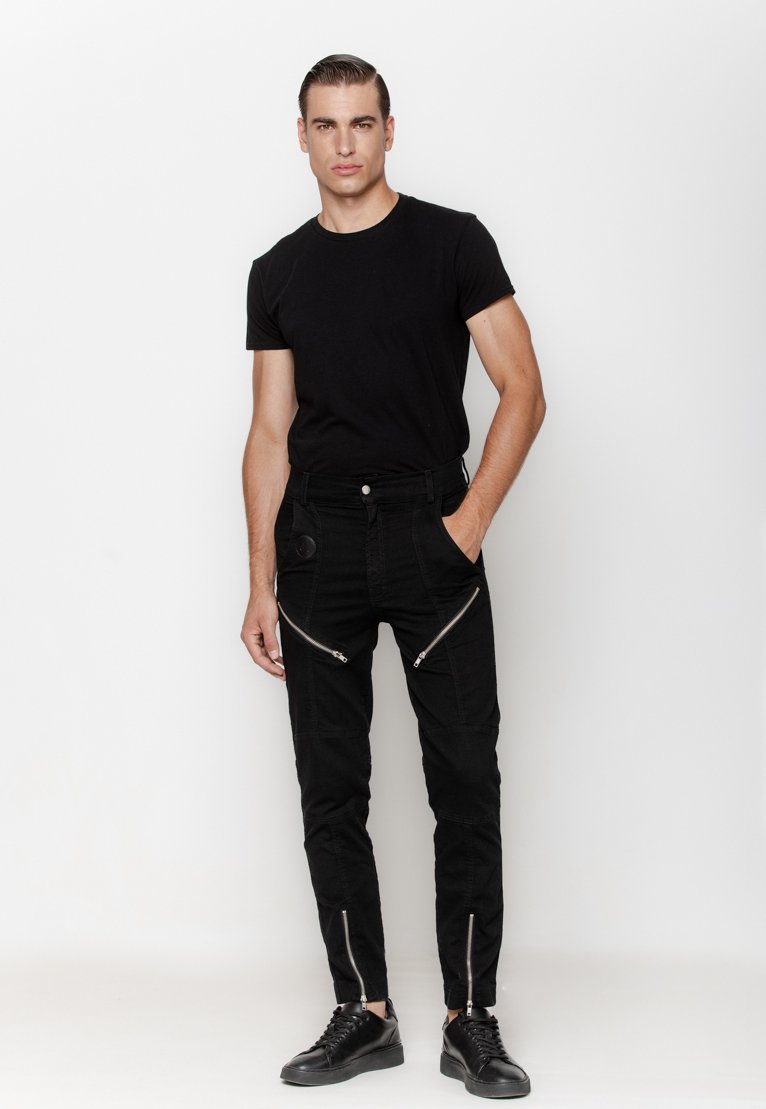 Czarny dopasowany t-shirt w połączeniu z czarnymi dżinsami z wieloma zasuwanymi kieszeniami i teksturowanymi panelami. Czarne sneakersy dopełniają outfit.