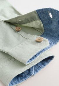 Light green shirt cuff with wooden buttons showing blue chambray fabric lining and buttonholes on a white background.