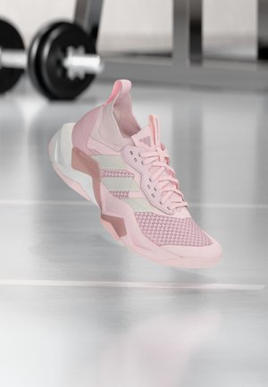 Világos rózsaszín Adidas sportcipő hálós felsőrésszel és fehér csíkokkal, egy edzőterem padlója felett lebegve, homályos kézi súlyzóval a háttérben.