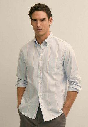 Młody mężczyzna o ciemnych włosach ubrany jest w jasnoniebieską koszulę na guziki z subtelnymi paskami, z podwiniętymi rękawami i rękami w kieszeniach, na neutralnym tle.