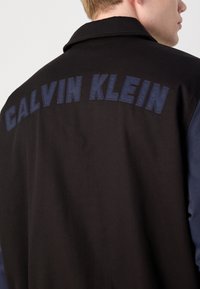 Black jacket with navy sleeves, featuring "CALVIN KLEIN" in large embroidered letters on the back. Smooth texture, collared design.