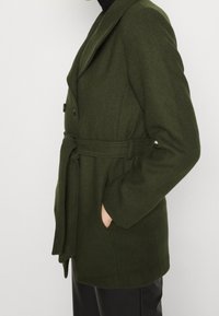 Abrigo verde oliva hecho de tejido texturizado, con cuello de chal, botones dobles, cinta en la cintura y bolsillos laterales.