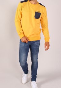 Camicia gialla a maniche lunghe con parte superiore textured, tasca blu scuro e polsini a coste; abbinata a jeans blu aderenti e sneakers bianche.