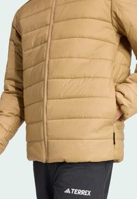 En person iført en beige puffjakke med lynlås foran står med den ene hånd i lommen, iført mørke bukser med et logo.