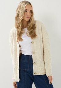 Cardigan beige con texture in un tessuto morbido e peloso; presenta grandi bottoni, polsini a coste e una tasca sul davanti. Indossato sopra una camicia bianca.