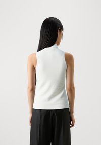 Short sleeveless top in white, featuring a high neckline and a smooth, textured fabric. View from the back. Paired with dark trousers.