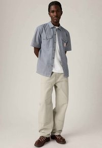 Camisa de manga curta azul e branca às riscas com dois bolsos no peito, combinada com calças cinza claras e sapatos castanhos com atacadores.