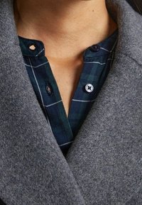 Abrigo de lana gris con solapas anchas, superpuesto a una camiseta de cuadros verdes. La camiseta presenta botones y detalles de costura visibles.