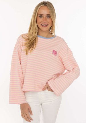 Junge Frau mit langen blonden Haaren lächelt, trägt ein rosa und beiges gestreiftes Langarmshirt und weiße Hosen, steht vor einem einfarbigen Hintergrund.