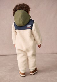 Fleeceový komplet v krémovej farbe s olivovo zelenou kapucňou a námorníckym horným panelom. Obsahuje logo a rebrované manžety. Viditeľná obuv dieťaťa.