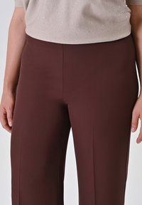 Donna che indossa pantaloni attillati marrone scuro e un maglione beige chiaro con leggere luccichii, in piedi con le mani rilassate lungo i fianchi.