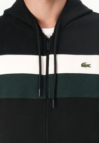 Crna dukserica sa zipom i dizajnom horizontalnih pruga u zelenoj i bijeloj boji. Ima mali izvezeni logotip krokodila i kapuljaču s dugmicama.