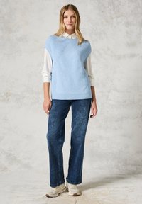 Jasnoniebieski dzianinowy sweter bez rękawów z wzorem w diamenty, noszony na białej koszuli z kołnierzykiem, w połączeniu z ciemnoniebieskimi, szerokimi dżinsami i trampkami.