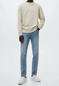 Persona che indossa una felpa beige, jeans azzurri chiari e sneakers bianche, in piedi con una mano in tasca contro uno sfondo semplice.
