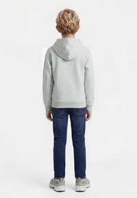 Copil cu păr blond și creț, văzut din spate, purtând un hanorac gri deschis, blugi albaștri închiși și adidași gri pe fundal alb.