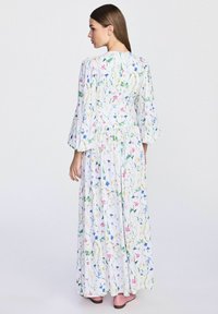 Długa, biała sukienka w kwiaty z marszczoną talią, długimi bufiastymi rękawami i wielokolorowymi wzorami kwiatowymi. Nosi z różowymi butami.