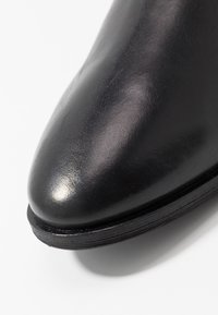 Närbild av en polerad svart lädersko med rundad tå och synlig sulkant på vit bakgrund.