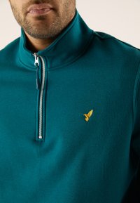 Suéter azul verdoso con cremallera hecho de tela suave. Presenta un cuello alto, cremallera plateada y un pequeño logo de pájaro dorado en el pecho.
