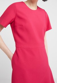 Sukienka z krótkim rękawem w jasnoróżowym kolorze, z okrągłym dekoltem, boczną kieszenią i widocznymi szwami wokół talii i dołu.