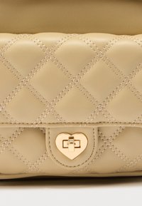 Μπεζ δερμάτινη τσάντα με ραμμένη επένδυση και κούμπωμα σε σχήμα καρδιάς από χρυσό, με διαγώνιες ραφές και απαλή υφή. Συμπαγές ορθογώνιο σχήμα.