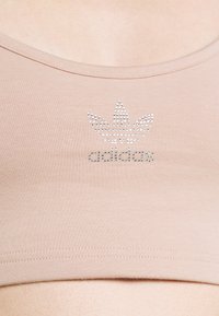 Bež sport bras izrađen od glatkog materijala s okruglim izrezom, s metalnim zakovicama u obliku logotipa koji je centriran na prednjem dijelu.