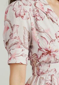 Abito floreale con uno sfondo chiaro e petali rosa e bordeaux; maniche a palloncino corte; vita con cintura; tessuto leggero con una texture morbida.