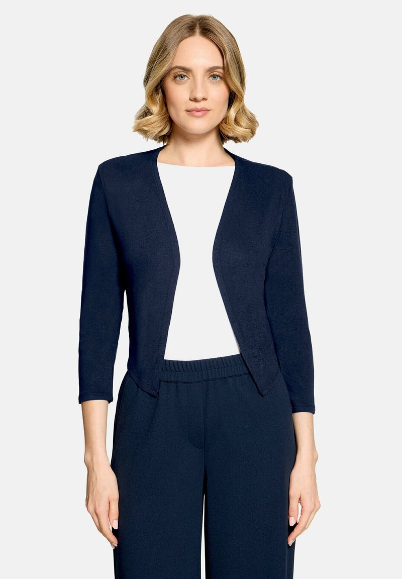 Femme aux cheveux blonds portant un cardigan bleu marine ouvert sur le devant, un haut blanc et un pantalon bleu marine avec taille élastique, debout devant un fond uni.