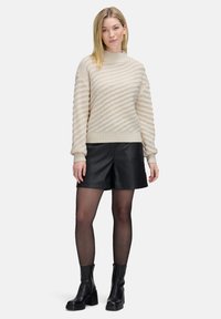 Frau mit glattem blonden Haar, die einen strukturierten beigen Pullover, schwarze Leder-Shorts, transparente Strumpfhosen und schwarze Knöchelstiefel trägt und vor einem weißen Hintergrund steht.