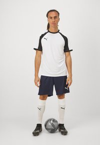 Camisola desportiva branca com detalhes pretos, calções azul-marinho, meias brancas, chuteiras pretas, de pé ao lado de uma bola de futebol preta e cinza sobre um fundo liso.