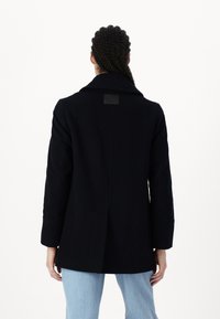 Abrigo de mezcla de lana azul marino oscuro con cuello de muesca, bolsillos laterales y una silueta elegante. La parte posterior presenta una etiqueta cuadrada en el cuello.
