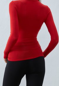 Rød langærmet top med en tætsiddende pasform, lavet af strækstof. Har en rund halsudskæring og en glat tekstur, båret med sorte leggings.