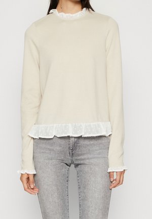 Femme portant un haut crème à manches longues avec un col, des poignets et un ourlet blancs volantés, associé à un jean gris clair délavé.