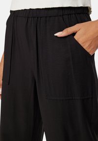 Zwarte broek van een gladde stof, met een brede elastische tailleband en zijzakken. Het ontwerp bevat plooien bij het zakgebied.