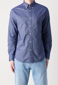 Tummansininen nappikauluspaita puuvillasekoituksesta, jossa on pieni logo vasemmassa rinnassa ja pitkät hihat, yhdistettynä vaaleansinihin farkkuihin.