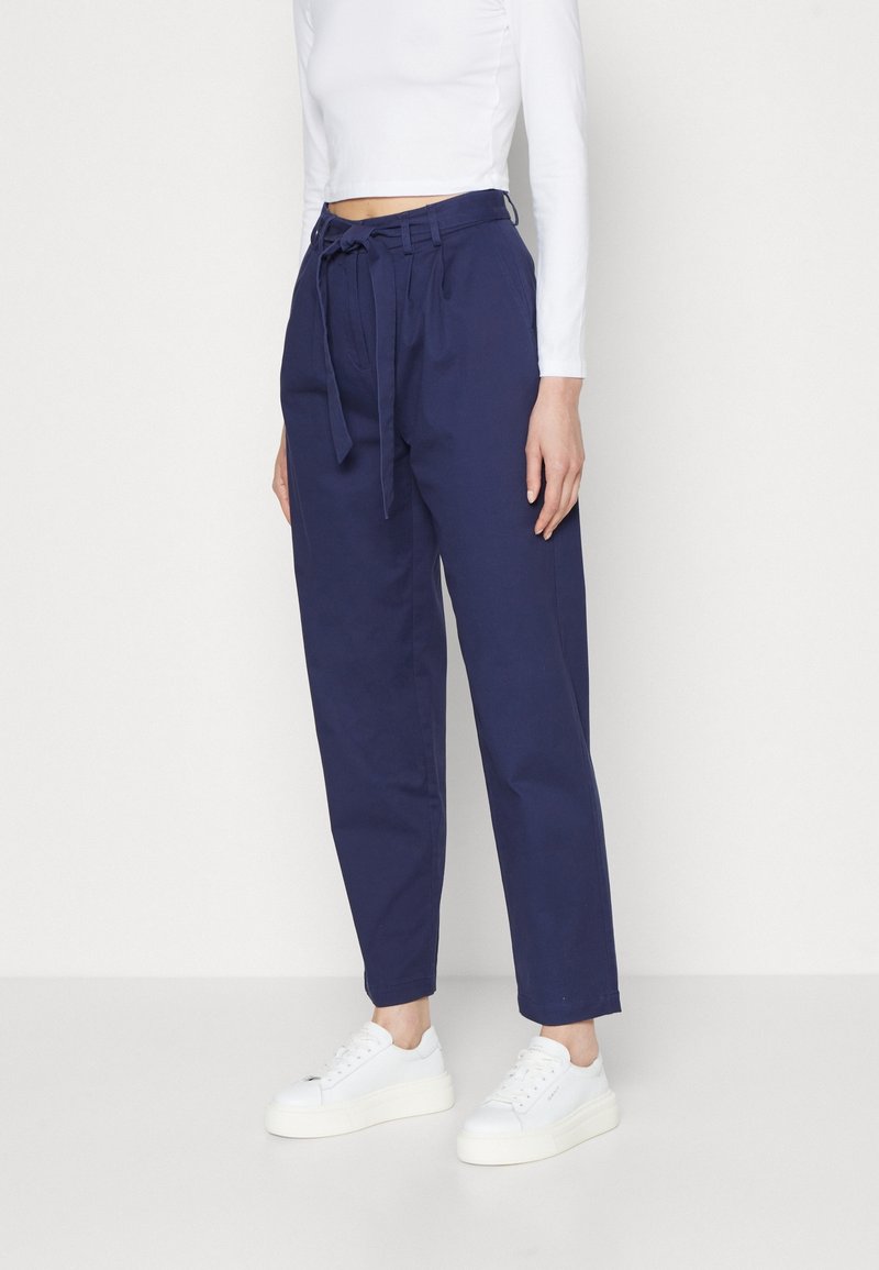 Pantalon en coton bleu marine avec une taille haute ceinturée, des jambes effilées et des poches latérales, associé à des baskets blanches. Texture lisse.