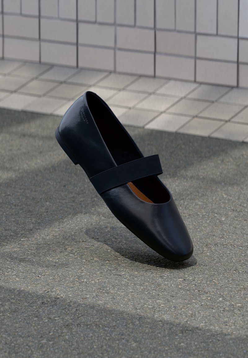 Chaussure noire style Mary Jane avec une bride, montrée légèrement en suspension au-dessus d’une surface en asphalte grise près d’un mur carrelé.