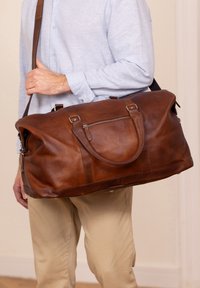 Sac de sport en cuir marron avec deux poignées et une bandoulière amovible ; comporte une poche zippée à l'avant. Texture lisse, design spacieux.