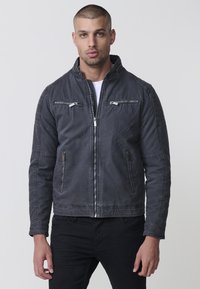 Férfi szürke farmerdzseki két cipzáras mellzsebbel, oldalsó zsebekkel és álló gallérral. Varrott részletezés a vállakon és az ujjakon.