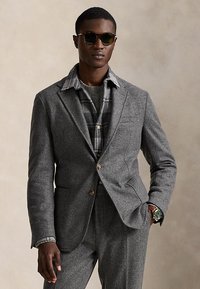 Άνδρας με σκούρα γυαλιά ηλίου, γκρι μάλλινο κοστούμι, πουκάμισο με καρό σχέδιο και ρολόι χειρός με πράσινο καντράν, ποζάροντας με το ένα χέρι στην τσέπη.