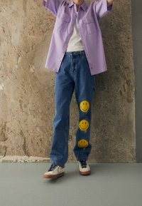 Ljuslila kortärmad skjorta, vit t-shirt under, blå jeans med tre gula smiley-ansiktstryck, och svarta sneakers med bruna sulor.