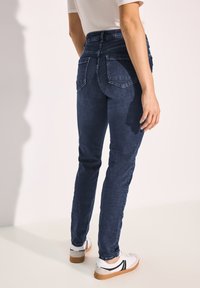 Jeans skinny bleu foncé en denim avec une taille mi-haute, dotés de deux poches arrière et d'une texture légèrement délavée. Portés avec des chaussures blanches.