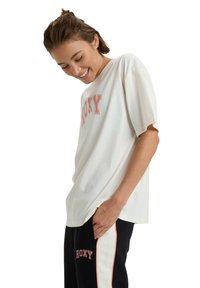 Mladá žena se usmívá, má na sobě bílý oversized tričko a černé kalhoty s bílými a oranžovými pruhy, oboje s logem "ROXY".